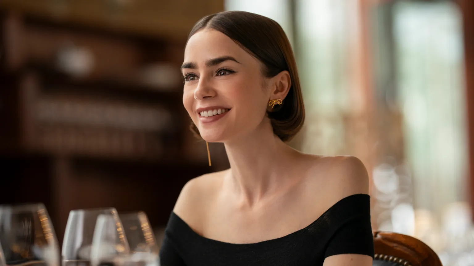 Caroline Dubois/Netflix A production still image of Lily Collins as Emily, pictured smiling in a restaurant. She has short brown bobbed air, and wears an off-the-shoulder black top and gold earrings.